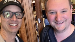 The top half of two men smiling for a selfie. The image is a bit blurry. One of the men is wearing a Ministry of Testing cap. There is wooden door just behind them and a corridor leading to a confe... image