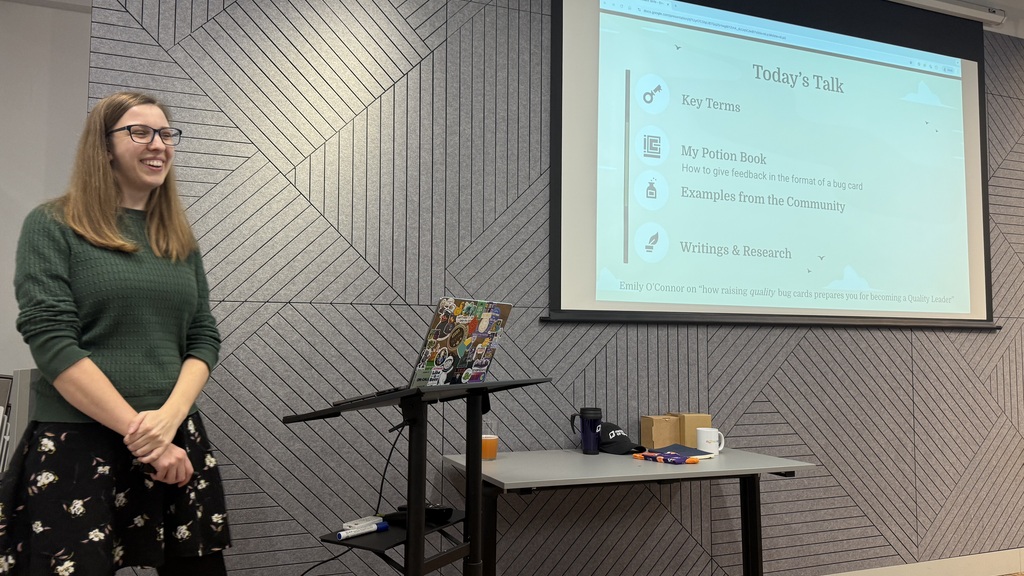 Emily stands to the side of a projector screen, wearing a green jumper and black skirt with flowers on. The screen shows a slide with today’s talk includes key terms, my potion book, how to give feedback, examples from the community, and writings and research 