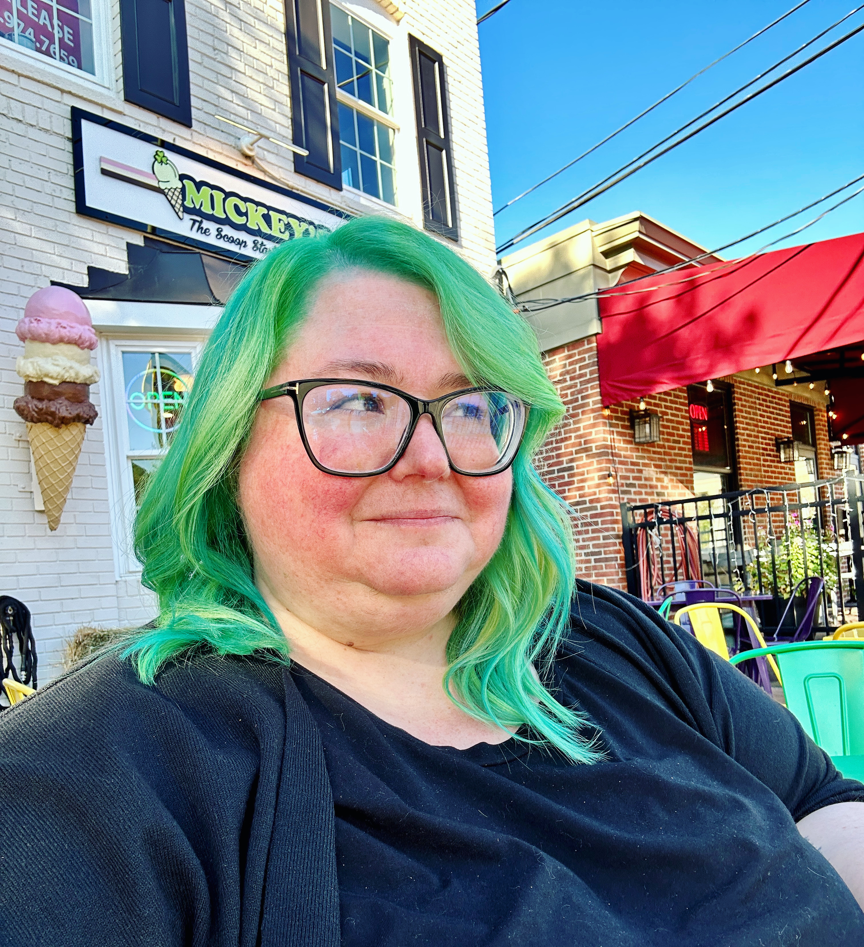 A nb person with green hair and glasses sits outside a restaurant with a sign that says Mickeys and an ice cream cone decoration on the wall. They are wearing a black shirt and sweater, and colorful chairs are visible nearby. They have green hair and are looking off into the distance