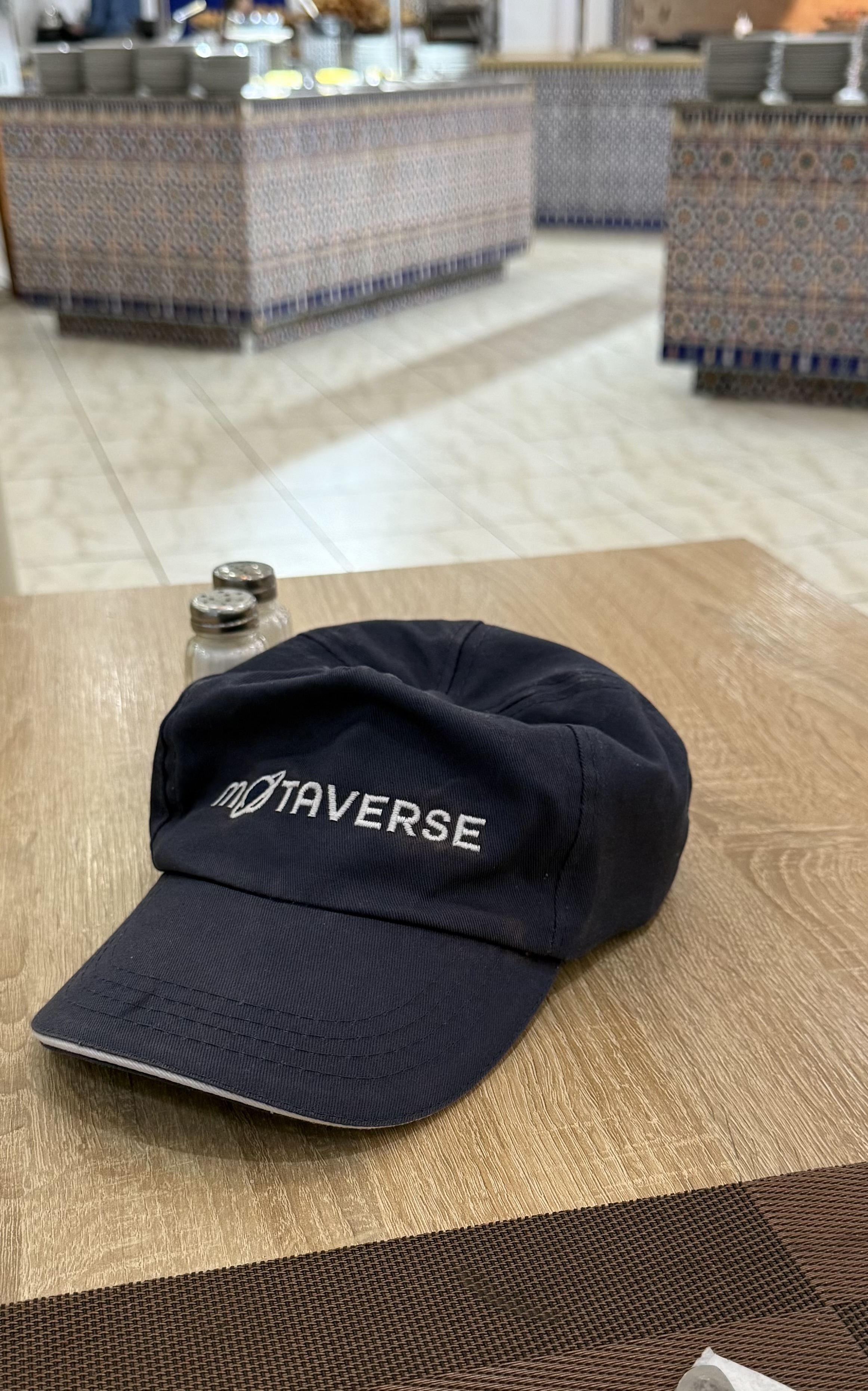 A blue baseball cap sits on a table with MoTaverse on it. In the background are serving tables with breakfast items on. 