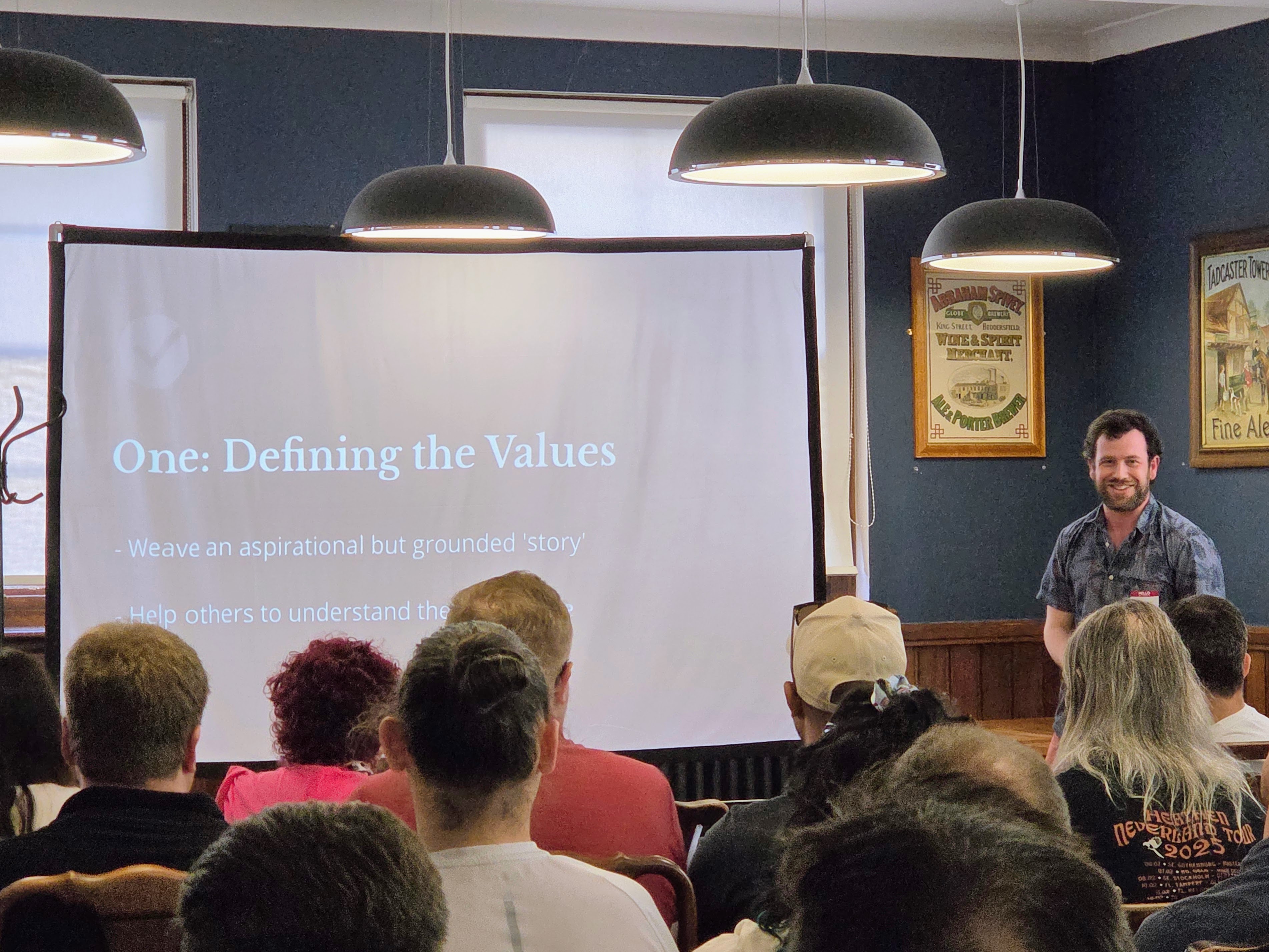 Chris Briggs giving a talk titled “The Good, The Right, and The Fitting” at Leeds Testing Atelier, standing beside a slide that reads “One: Defining the Values” in a room full of attendees.