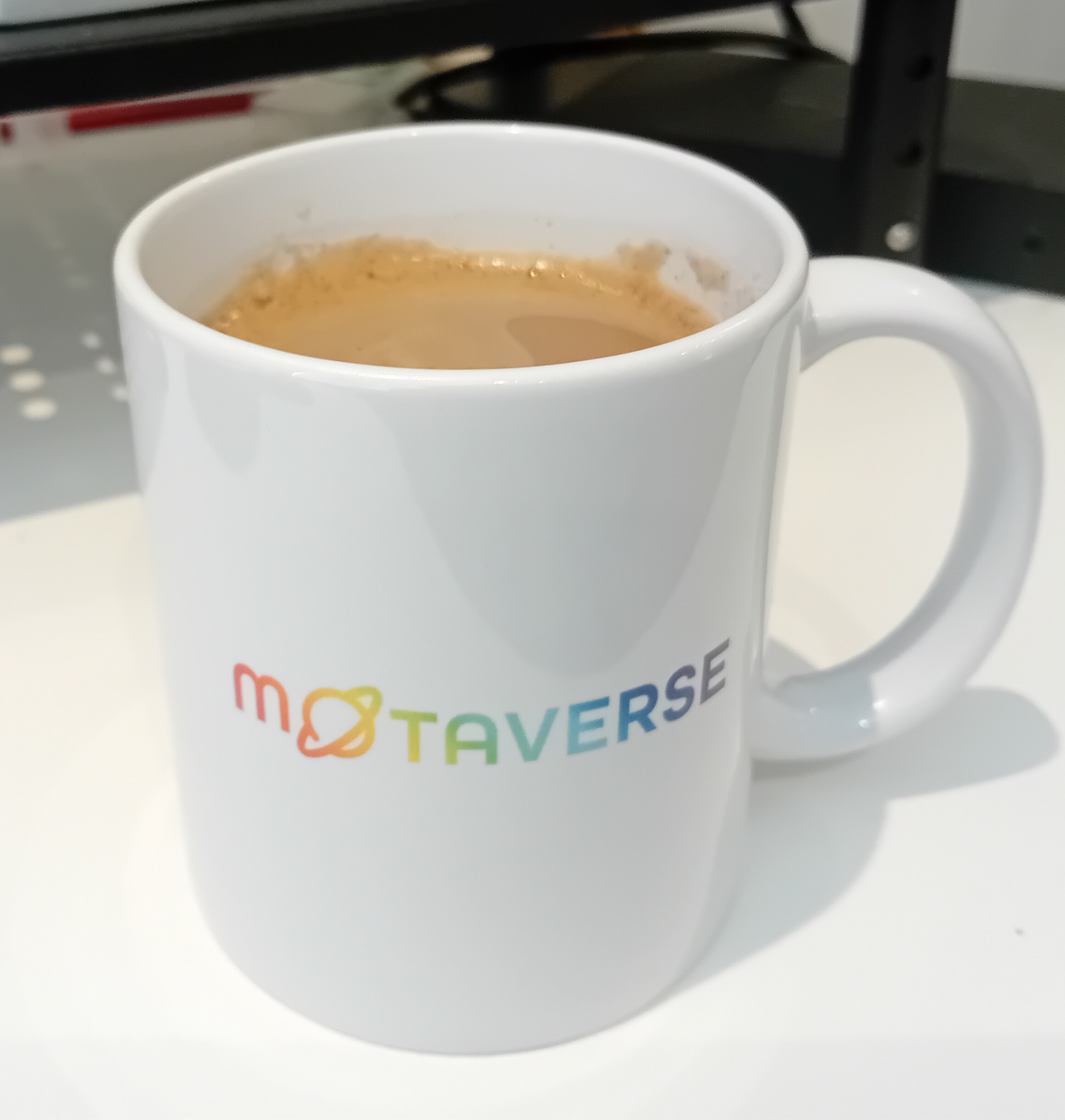 White coffee mug on a desk with multi coloured lettering spelling the word Motaverse.