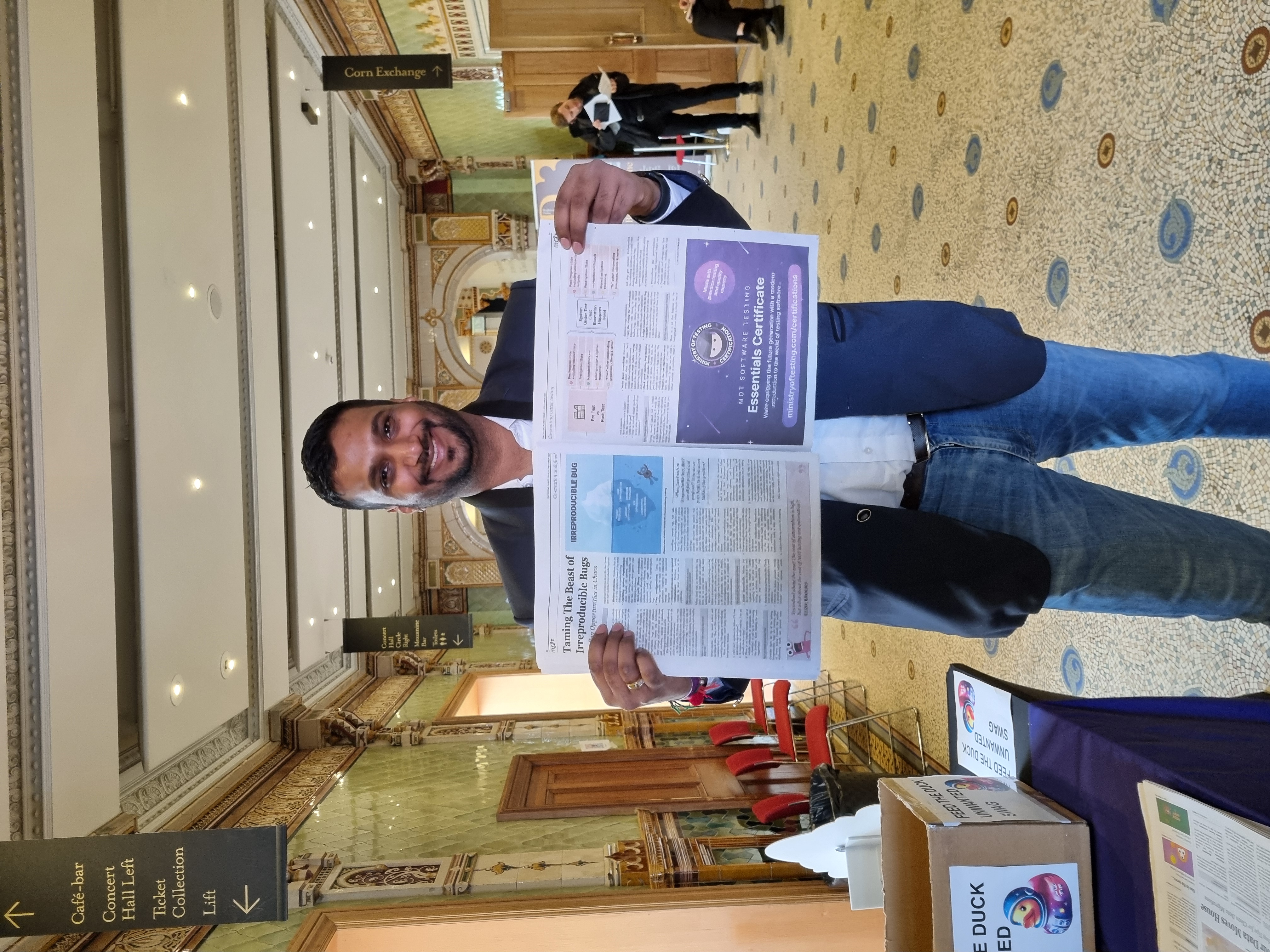Rahul Parwal stands proudly at TestBash 2024, holding up the latest edition of The Testing Planet newspaper with his article "Taming the Beast of Irreproducible Bugs" prominently featured. He smiles at the camera in the very grand venue, the Brighton Dome
