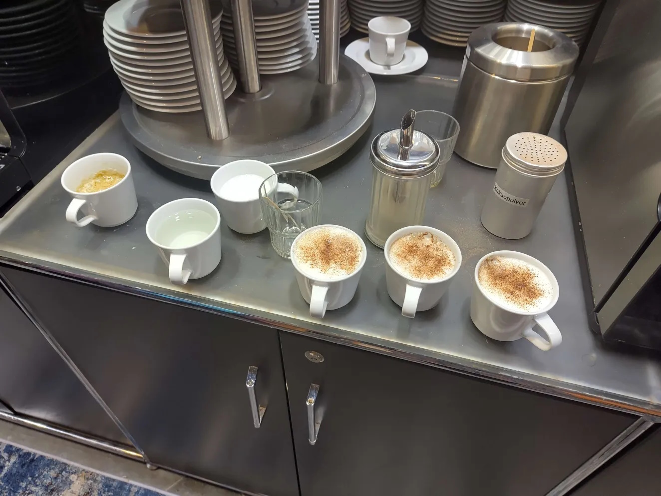 A countertop with several coffee cups, glasses, and containers arranged beside a stack of plates and two coffee machines. The cups in front are filled with different hot beverages. A sugar dispenser and a cocoa powder shaker are placed between the cups.