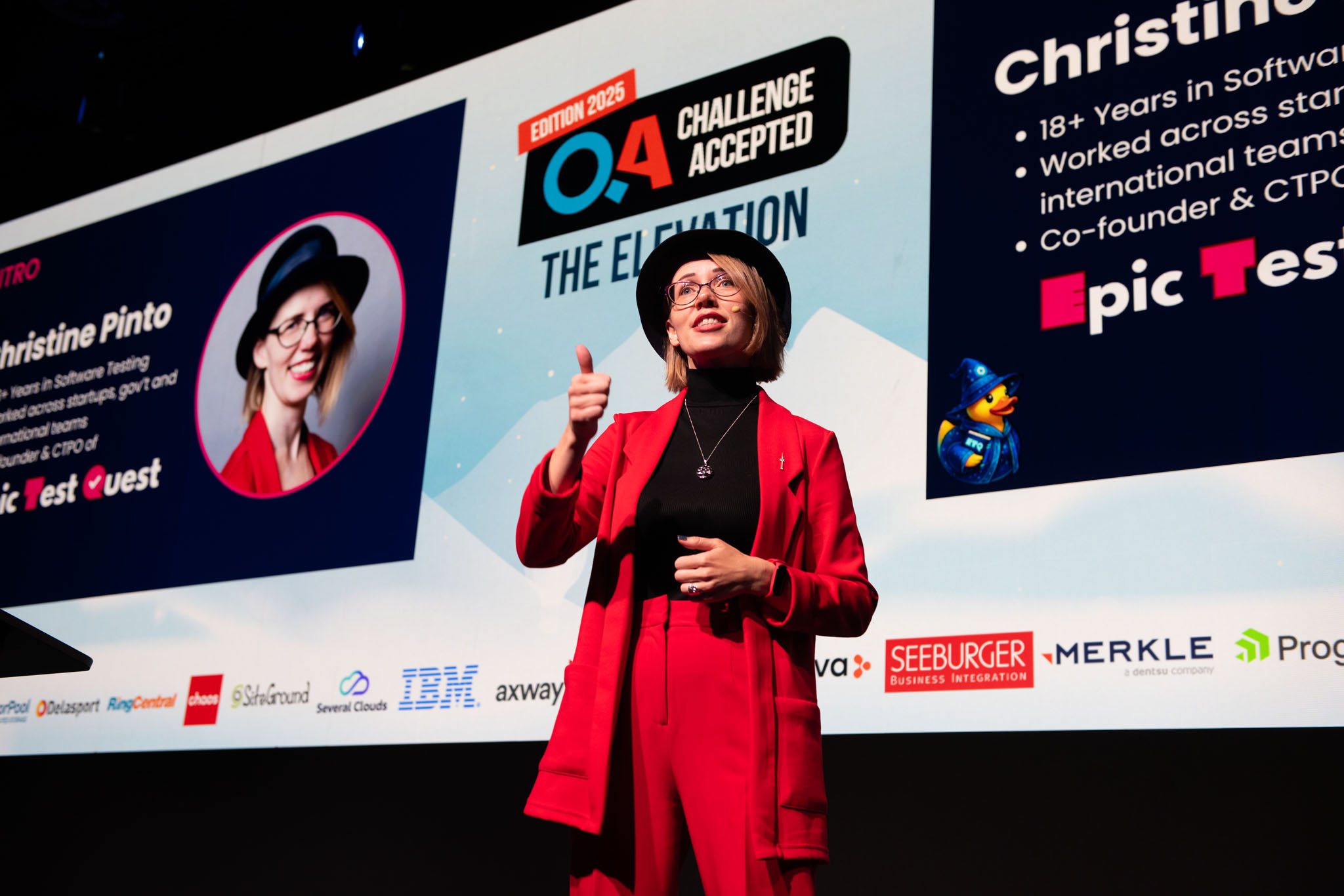 Christine Pinto speaking on stage at the QA: Challenge Accepted 2025 conference in Sofia, Bulgaria. She’s wearing a red suit and black hat, giving a thumbs-up while presenting Epic Test Quest, with her intro slide displayed behind her.