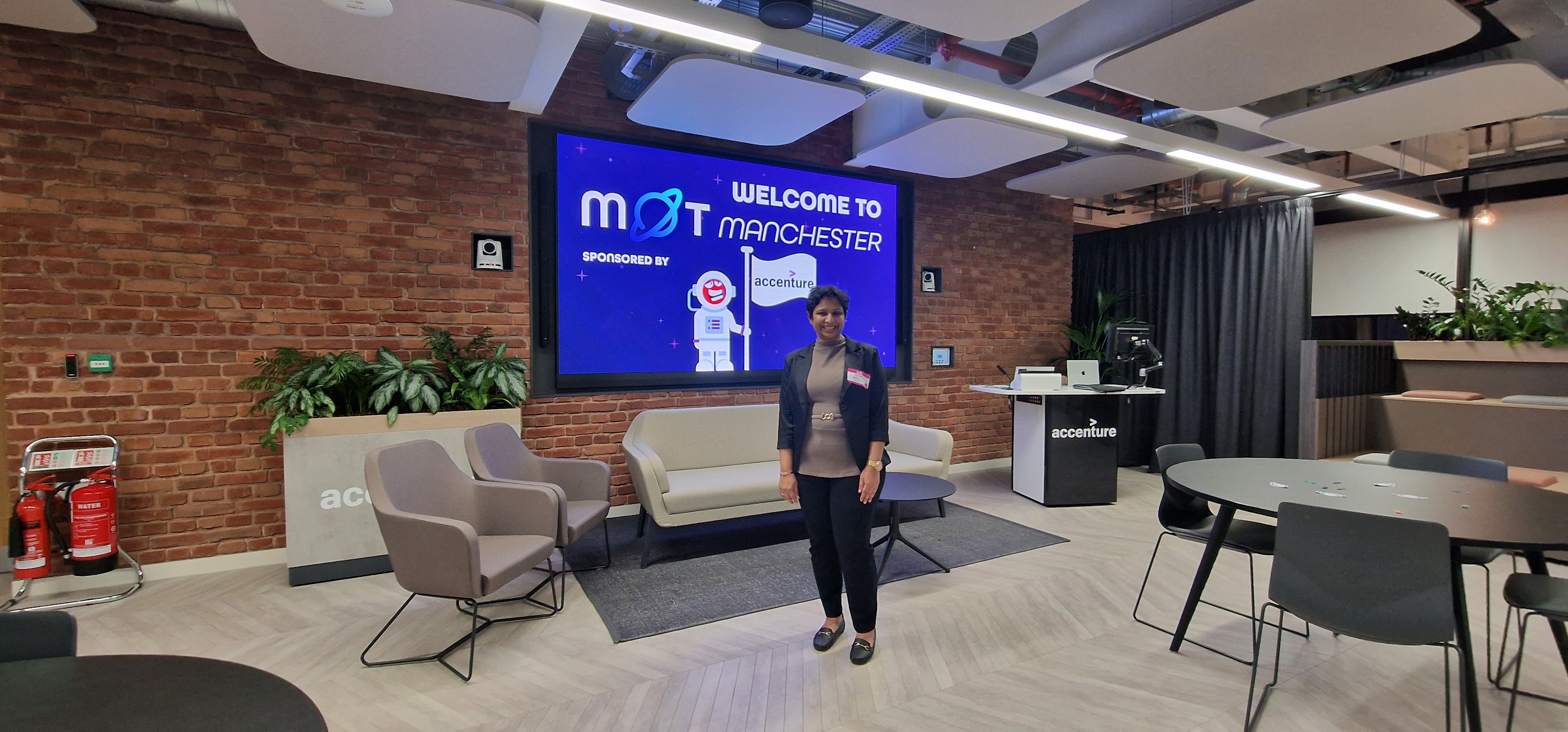 A person stands smiling in the center of a modern, open-plan office lounge. They are wearing a dark blazer over a tan... image