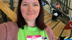 A selfie of Sarah K at her first TestBash in Brighton in 2019. She is wearing a shocking bright green MoT shirt standing by the main staircase in the venue that has MoT flags hung along the bannister. image
