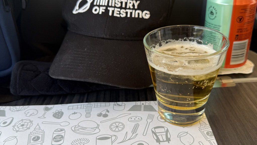 View from my seat of the first class mat, with a larger drink on it. A black ministry of testing cap is at the back.  image