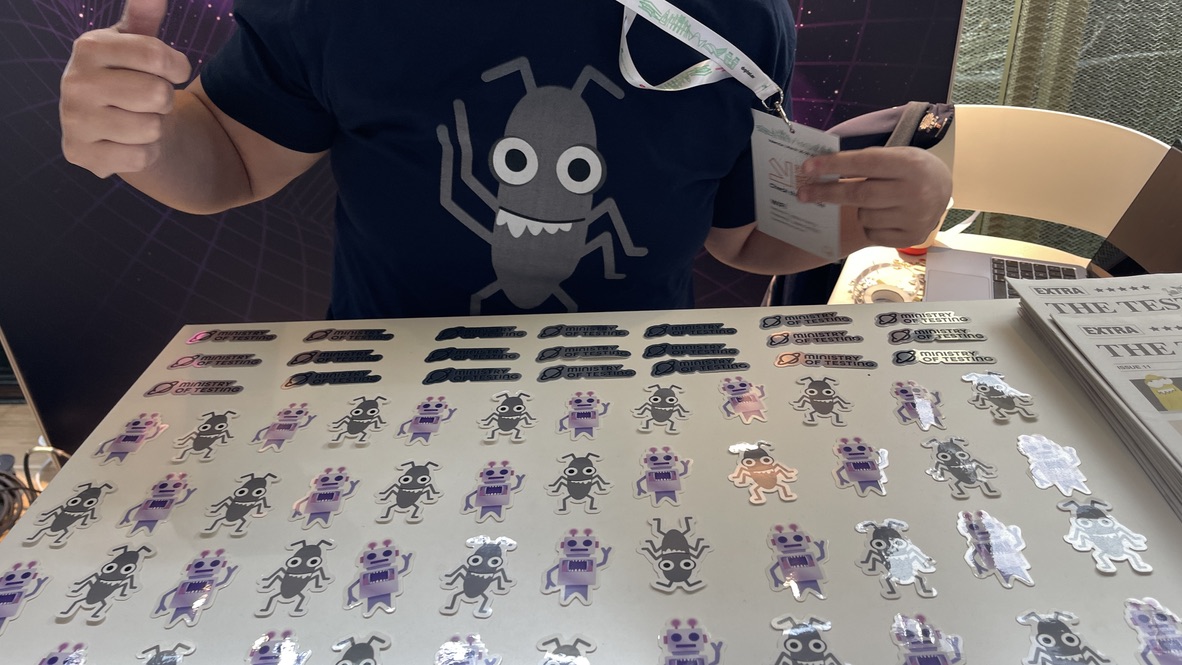 A close-up of the Ministry of Testing stand at Selenium Conf 2025, showing a person wearing the iconic MoT bug t-shirt and a display of colorful bug stickers, alongside stacks of The Testing Planet zines.