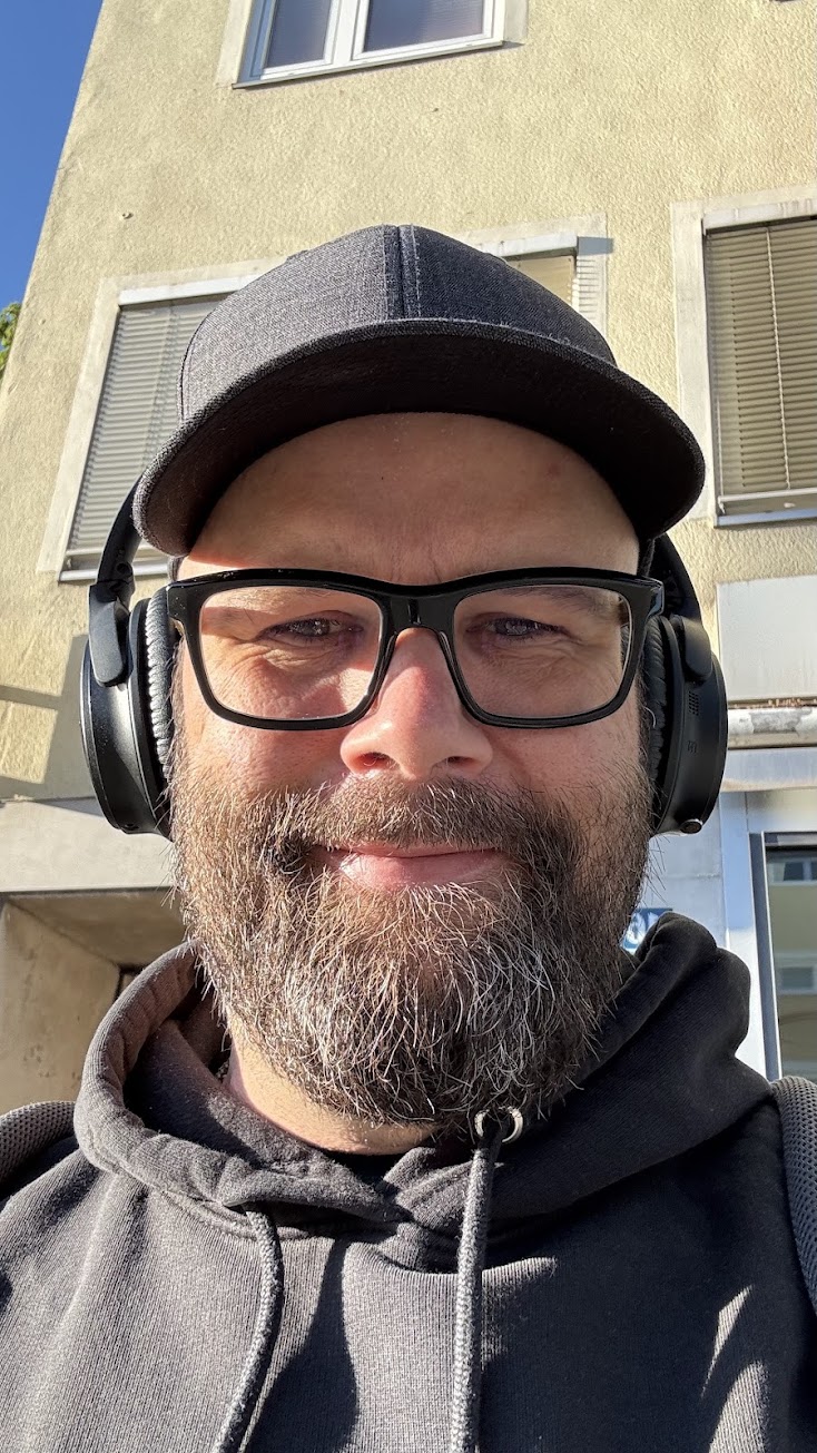 Bearded man wearing a dark cap and a light gray hoodie, standing outdoors.