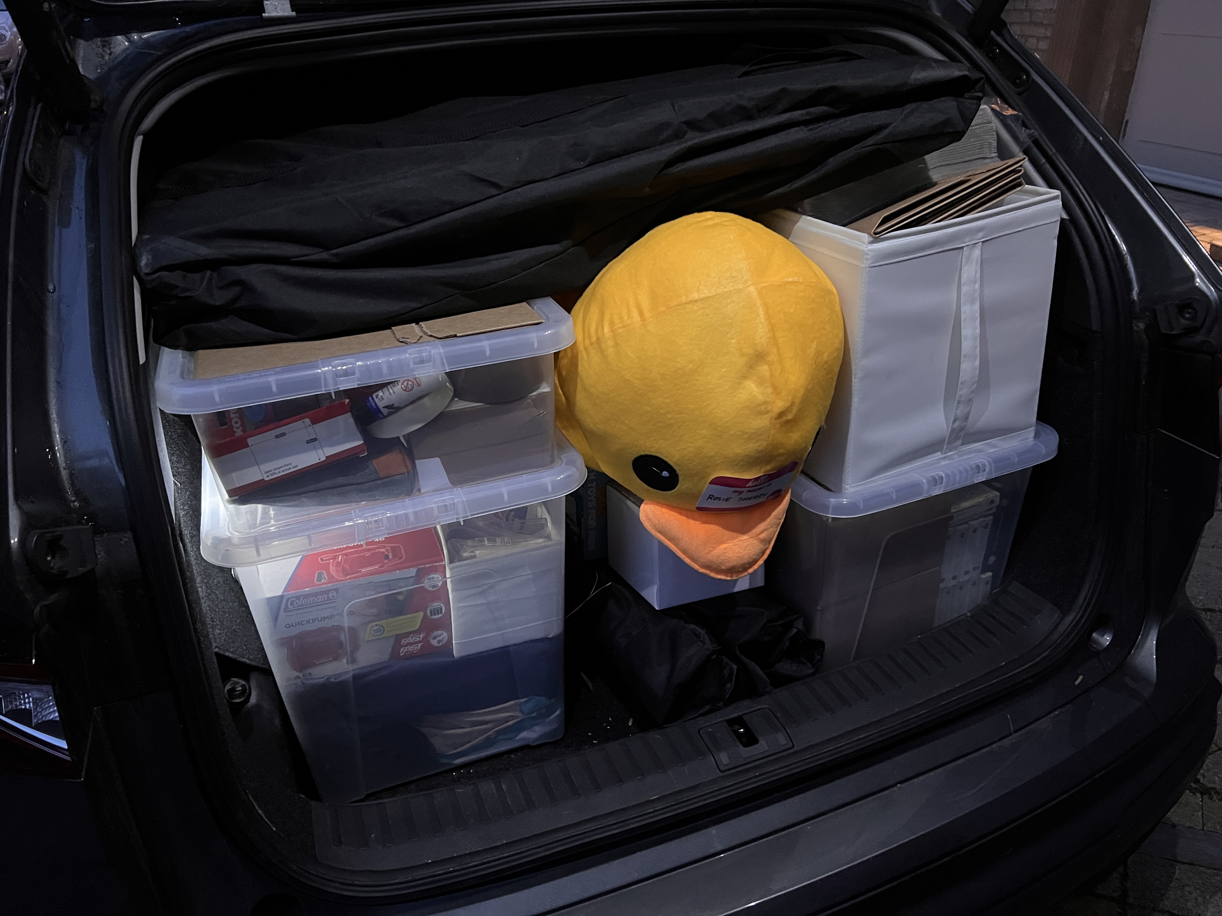 The boot of a large car. There is a big fluffy duck toy poking its head out next to three large plastic containers with stationary in them. There are other event items packed into the boot space. It's crammed tight. 