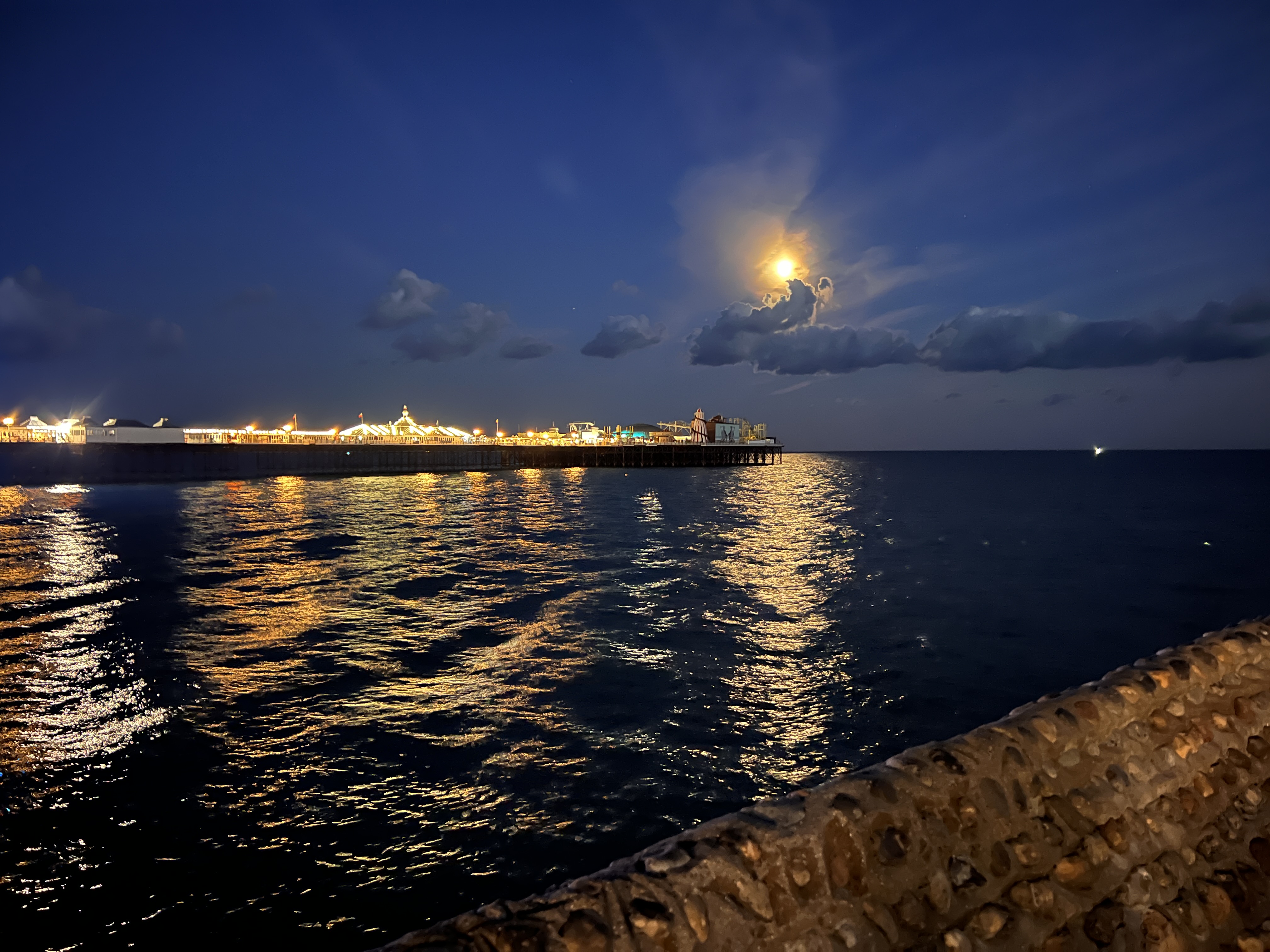 The sea at night. There is a lit-up pier. The moon shines through a small set of clouds and reflects its light on the wavey sea. 