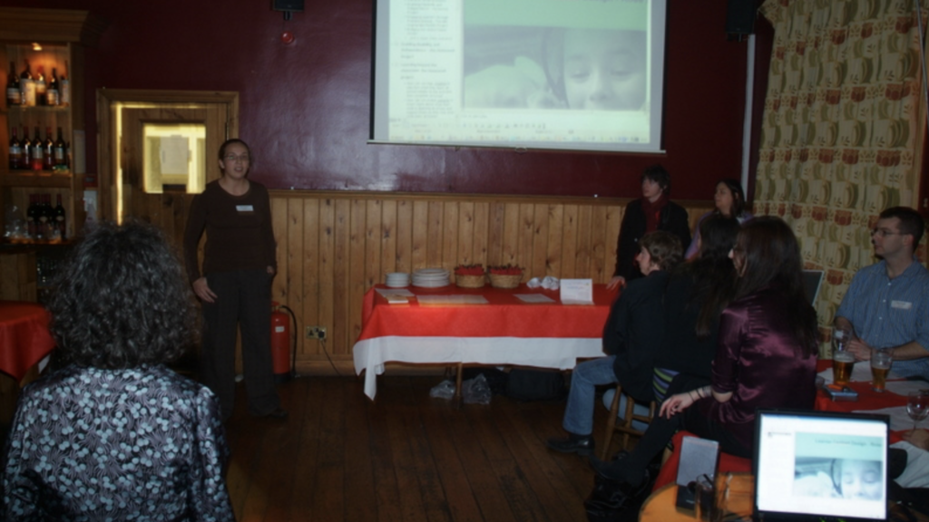 Rosie standing in the front of a room in a pub, saying something at what was a Girl Geek Dinner meetup. image