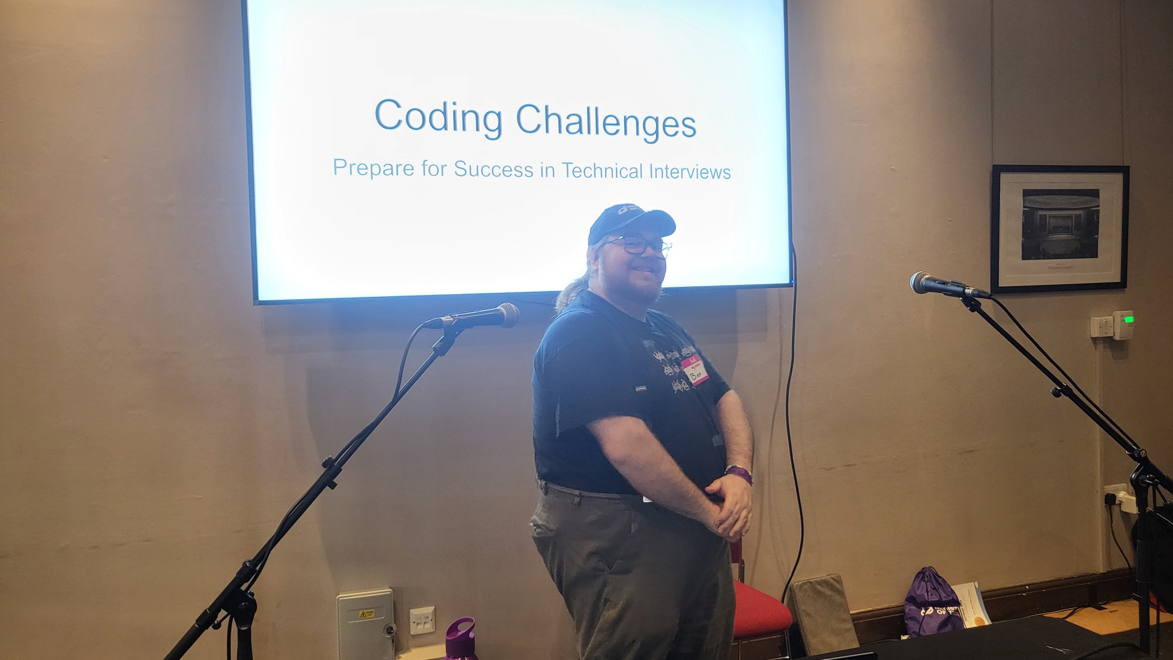 Ben Dowen, a bearded man wearing grey trousers a black short sleeved tshirt, pink name badge and black cap, stands proudly infront of his own title slide that reads "Coding Challenges Prepare for Success in Technical Interviews". 