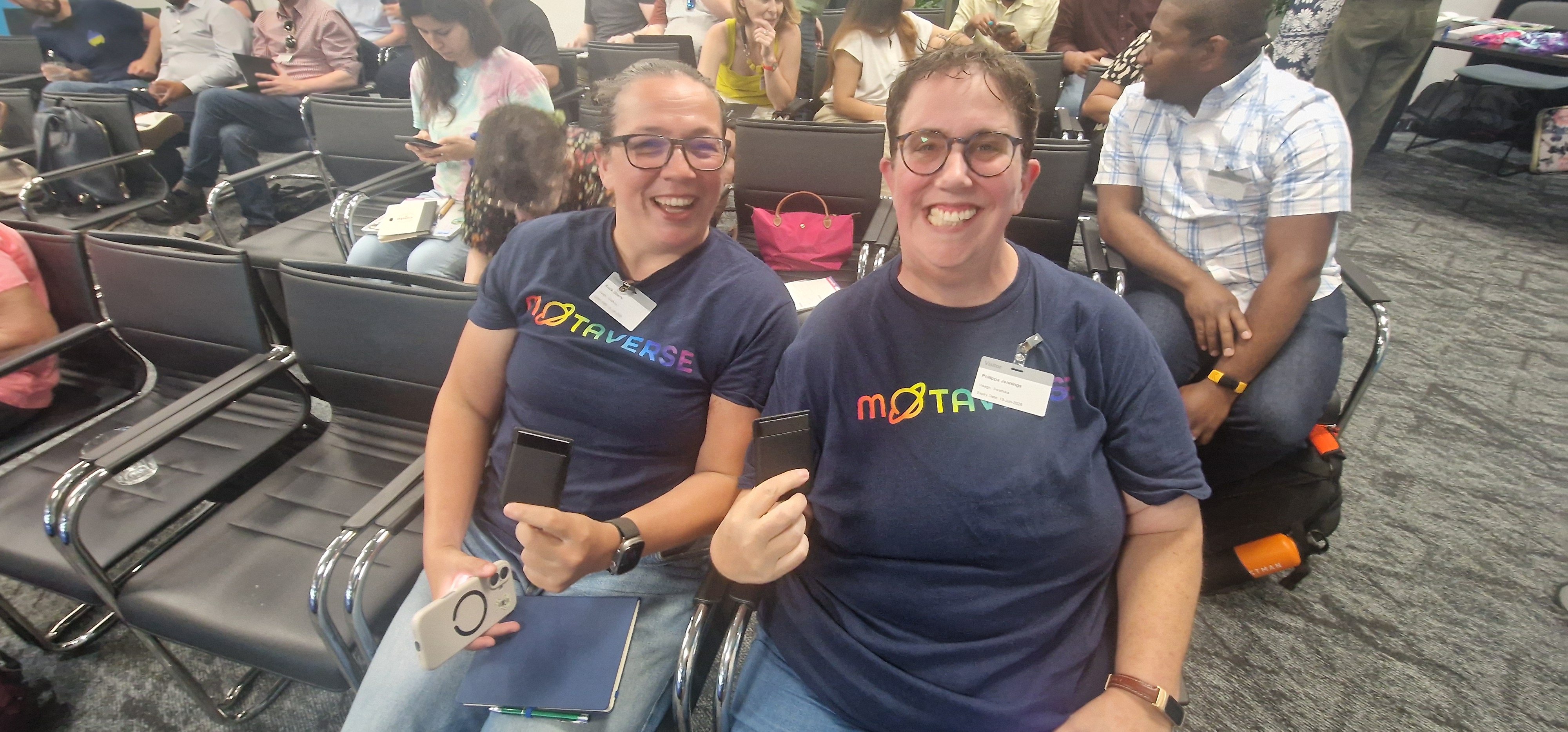Two smiling people wearing matching blue "METAVERSE" t-shirts sit in an auditorium-style room filled with black chair... image
