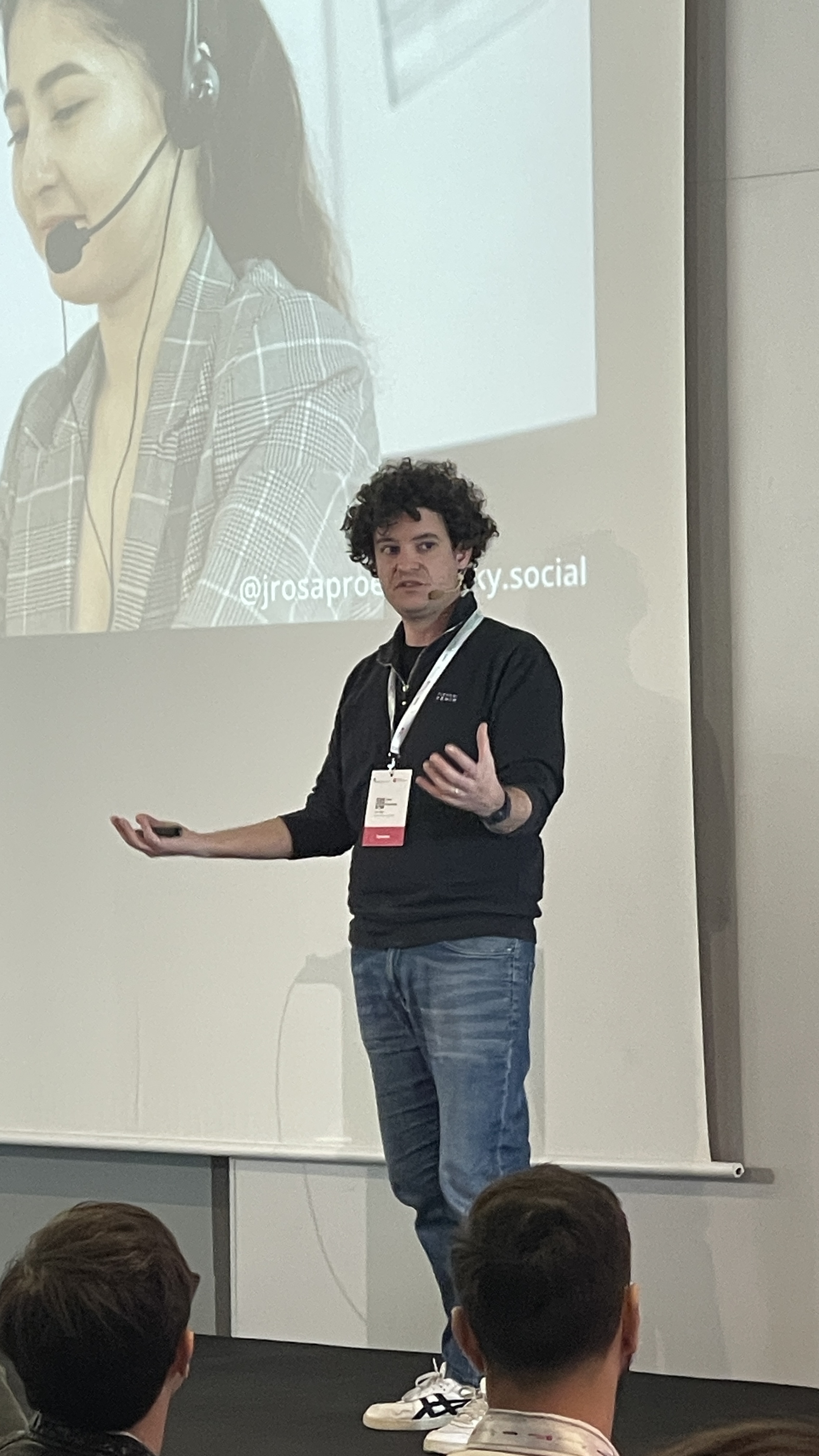 João Proença stands on stage mid-talk at Selenium Conf 2025, gesturing with energy while presenting. His talk is being projected behind him, and audience members are visible in the foreground.