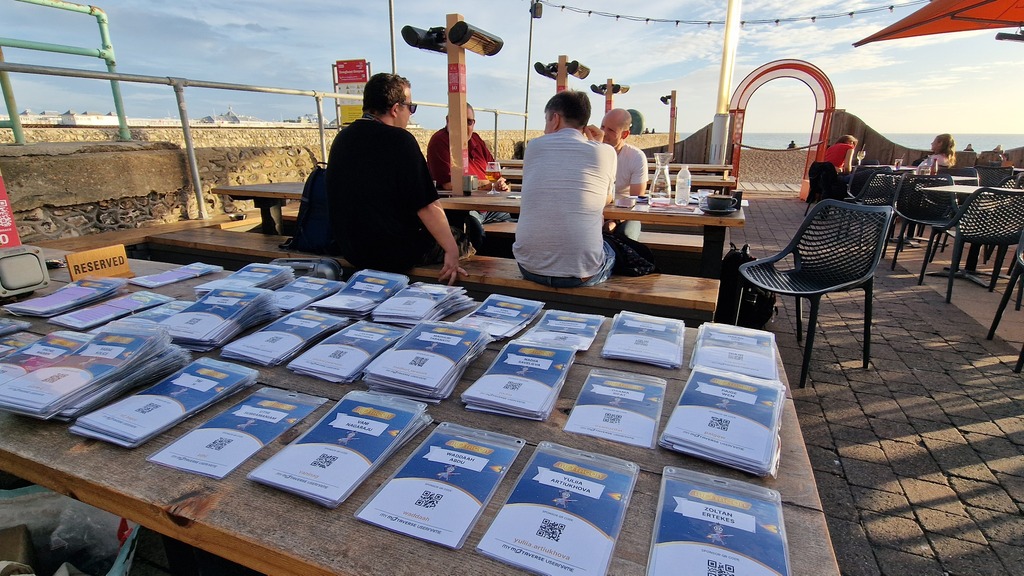 Image of a wooden table with a lot of conference passes layed out on top. A group of people sit on a table behind.  image