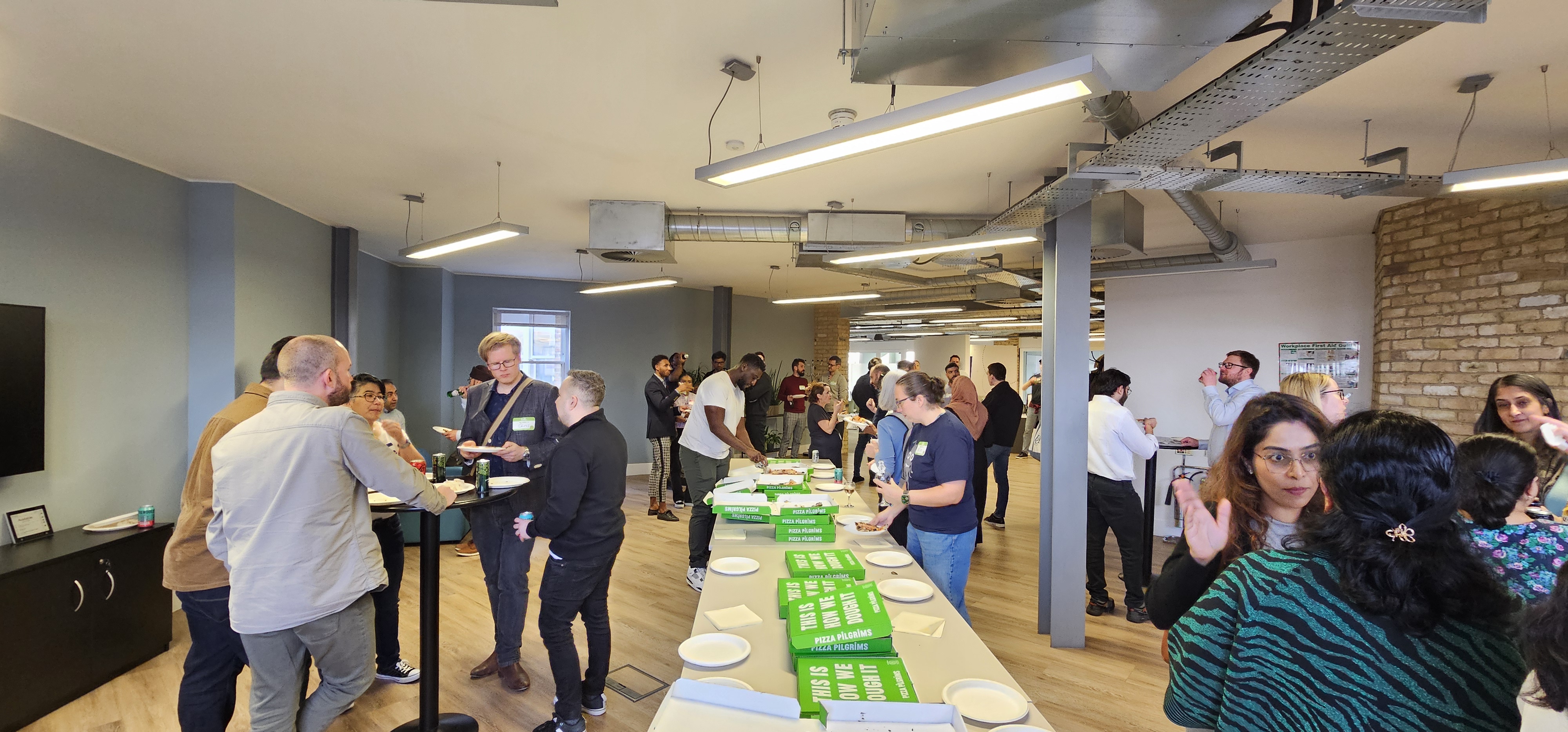 A large group of people conversing, eating and drinking in a room.

There is a long table with green pizza boxes and paper plates 