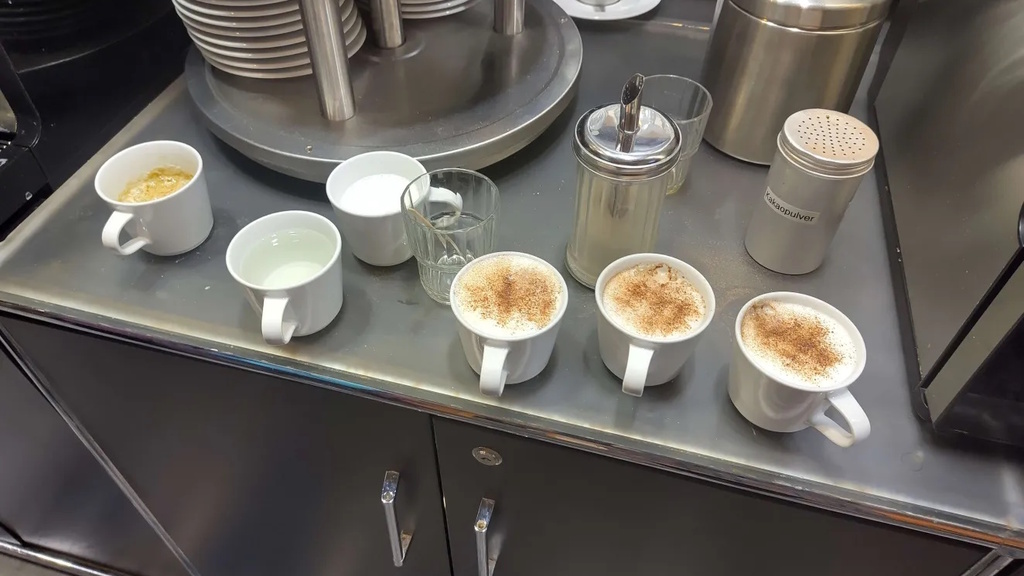 A countertop with several coffee cups, glasses, and containers arranged beside a stack of plates and two coffee machines. The cups in front are filled with different hot beverages. A sugar dispenser and a cocoa powder shaker are placed between the cups.